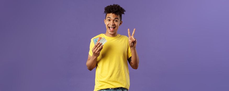 Health, influenza, covid-19 concept. Portrait of upbeat, healthy young man feel much better after taking vitamins or tablets, hold prescribed drugs, show peace sign, smiling pleased.