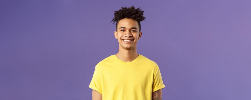 Close-up portrait of nice, friendly-looking hispanic male student in yellow t-shirt, grinning delighted, look upbeat happy and positive, standing enthusiastic with beaming smile purple background.