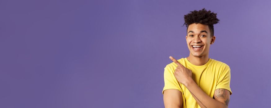 Close-up portrait of cheerful, attractive young male student showing promo, pointing at upper left corner, smiling pleased, recommend click link, visit page or download app, purple background.