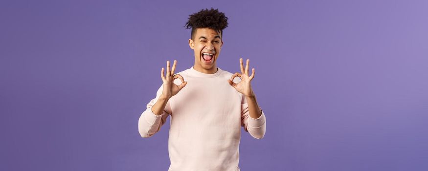 Portrait of chill young relaxed handsome man assure everything good, saying all okay, smiling and winking, recommend awesome product or online shop, leave positive feedback, purple background.