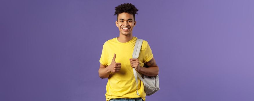 Back to school, university concept. Portrait of cheerful handsome male student describe his summer vacation to classmates, show recommendation, thumbs-up approval, hold backpack.