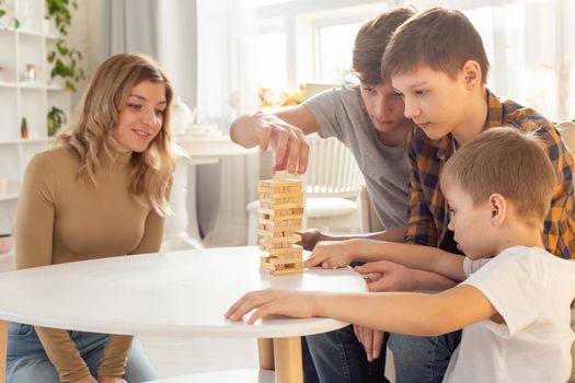 Family, three boys and woman, in a sunny room, are enthusiastically playing a board game made of wooden rectangular blocks, pulling pieces out of the tower. Close-up