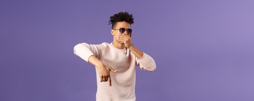 Waist-up portrait of cool young handsome guy with dreads, sunglasses on, hold hand over mouth while beatboxing, waving hand to shake audience up, reading own rap, purple background.