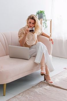 stylish woman in light clothes sitting on sofa, working on laptop, drink a cup of coffee. Vertical. Copy space