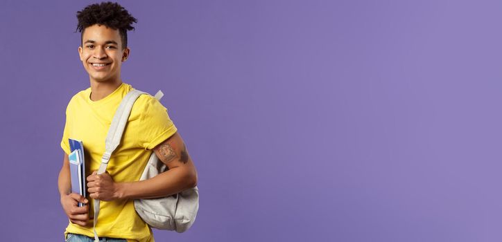 Back to school, university concept. Portrait of handsome charismatic hispanic guy, student walking to campus, going for lecture, studying hold backpack and notebooks, stand purple background.