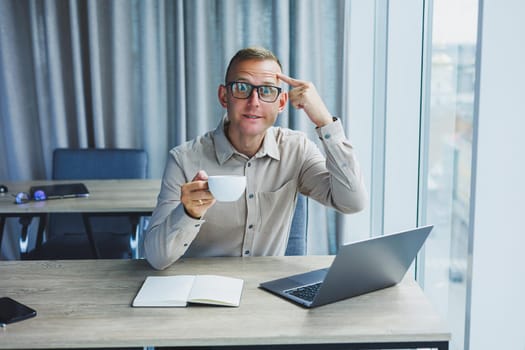 A freelancer in glasses works on a laptop and drinks coffee, a manager sits at a table in the office, works on a laptop. Freelancer workplace, employee at remote work.
