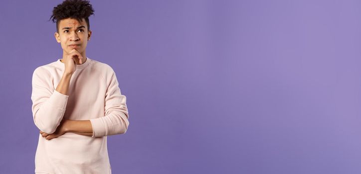 Portrait of complicated, young thoughtful man with dreads, look troubled up, thinking what to do, standing indecisive, grimacing not knowing answer, facing hard choices, purple background.