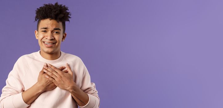 Close-up portrait of handsome silly hispanic guy with dreads, place hands on heart and sighing as contemplate something adorable speaking from all his soul, thanking for praises, purple background.