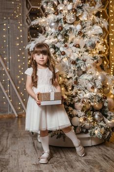 A girl in a festive light dress with a gift in her hands next to a Christmas tree, lights of garlands in the background. The concept of New Year holidays.