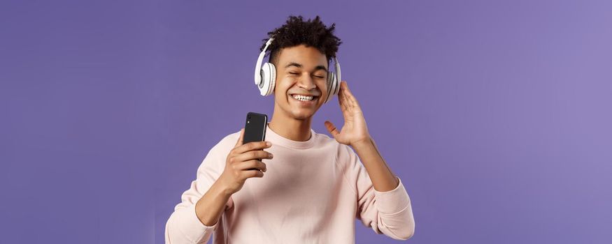 Technology and lifestyle concept. Portrait of carefree attractive young man enjoying fine sound, listening music with headphones look delighted awesome quality of bass, hold mobile phone.