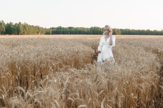 A blonde woman in a long white dress walks in a wheat field. The concept of a wedding and walking in nature.