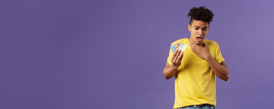 Health, influenza, covid-19 concept. Portrait of young man coughing, choking with tablet, drank pill without water, holding drugs and touch neck as suffer sore throat, purple background.