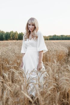 A blonde woman in a long white dress walks in a wheat field. The concept of a wedding and walking in nature.