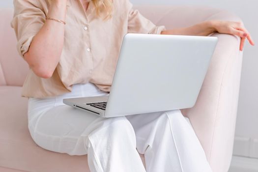 Business woman in light clothes, sitting on a light pink sofa, with a gray open laptop on her knees, in the office close-up