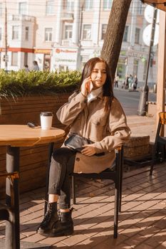 A stylish brunette woman is sitting at a table in an outdoor cafe with coffee and a phone. The girl is talking on the phone. Autumn walk.