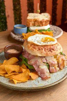 photo of delicious bacon burger accompanied with an egg, chipotle dressing and dehydrated bananas on wooden table. big hamburguer. accompanied with a panela sandwich