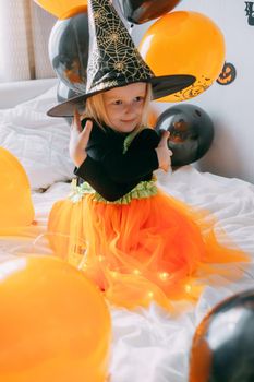 Children's Halloween - a girl in a witch hat and a carnival costume with airy orange and black balloons at home. Ready to celebrate Halloween.