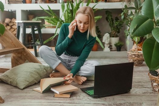A blonde woman in a room with a lot of green indoor plants is working on a laptop. The concept of biophysical design in the interior. Work from home, work as a freelancer.