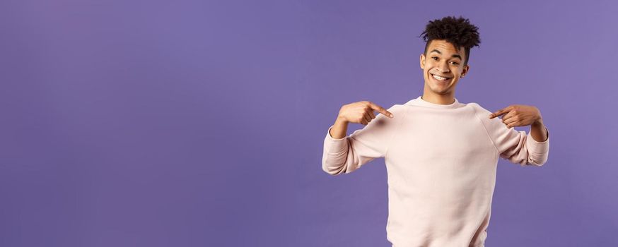 Cute and silly handsome male student pointing at himself and making lovely smile, ready to help offer himself, want participate or being chosen, volunteer to be a candidate, purple background.