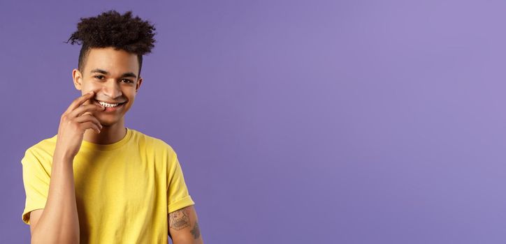 Close-up portrait of lovely young hispanic gay man with dreads, tattoos, touching lip sensually and flirty smiling, checking out someone really cute and handsome, standing purple background.