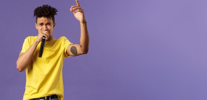 Portrait of young carefree sassy hipster guy with tattoos, yellow t-shirt performing in front audience with his hip-hop or rap, singing song, raising finger up, hold microphone, karaoke.