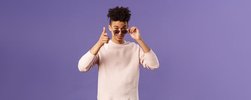 Portrait of cheeky handsome young man wearing sunglasses, take them off and show thumb-up, like or approval, recommendation sign, smirk satisfied, saying all good, alright, purple background.