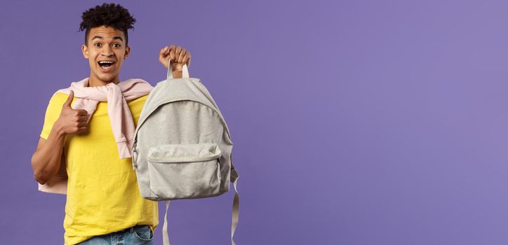 Portrait of cheerful young handsome male student, man recommending backpack, holding rucksack and show thumbs-up, bought new equipment for university semester, purple background.