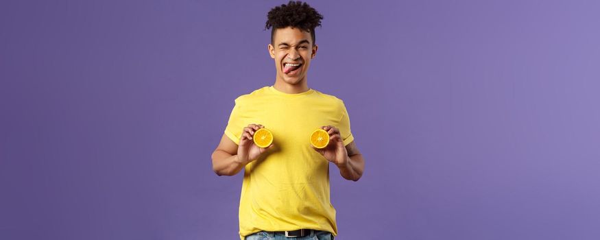 Holidays, vitamins and vacation concept. Portrait of sassy and cheeky funny young playful guy, fool around with fruit, holding pieces of oranges like women breast, show tongue smiling.