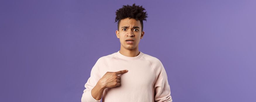 Close-up portrait of embarrassed and worried young man being named or called to principal office, pointing at himself and asking why with disbelief and anxiety, standing purple background.