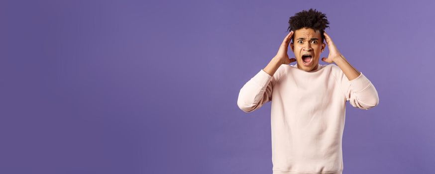 Portrait of scared and anxious young hispanic handsome man in panic, shaking hands nead head, screaming and gasping afraid, being in trouble, standing alarmed over purple background.