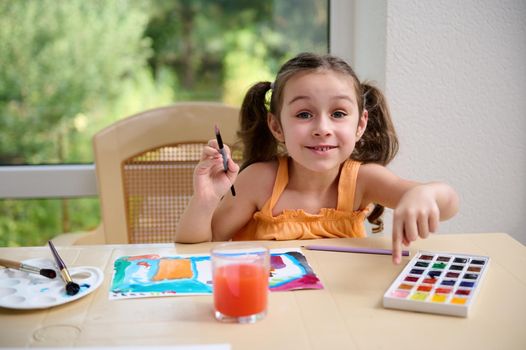 Happy little girl sitting at table and cutely smiling at camera while painting with watercolors and paintbrush. Kids education and development. Drawing. Art classes and courses for telented children