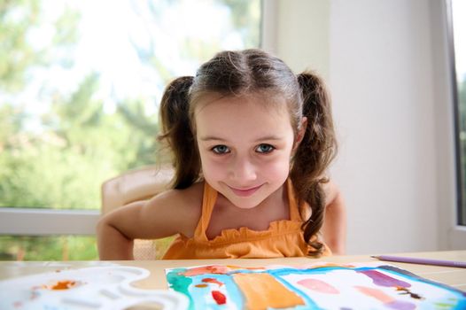 Cheerful European little girl with two funny ponytails wearing an orange top, sitting at a table with her drawn picture, during a watercolor painting art class and cutely smiling looking at the camera