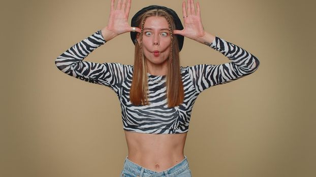 Lovely pretty funny woman in crop top making playful silly facial expressions and grimacing, fooling around, showing tongue. Adult stylish female girl isolated alone on beige studio background indoors