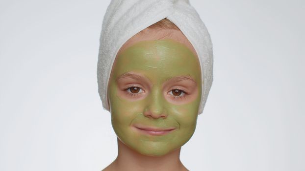 Smiling child girl in towel on head applying cleansing moisturizing green cucumber vitamin facial mask. Teenager kid face skin care treatment, natural cosmetics. Female portrait. Perfect fresh clean