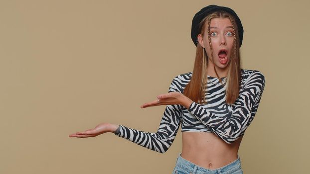 Lovely pretty young woman showing thumbs up and pointing left empty place, advertising area for commercial text, copy space for goods promotion. Adult stylish girl isolated on beige studio background
