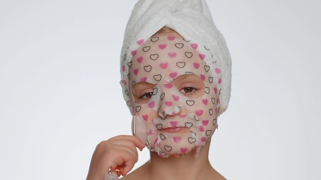 Beautiful young smiling child girl applying cosmetic moisturizing face mask, use massage roller. Teenager kid skin care treatment, natural cosmetics. Female close-up portrait on white background