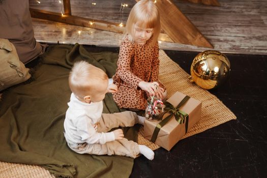 Little brother and sister play on Christmas eve in a beautiful house decorated for the New Year holidays. Children are playing with a Christmas gift. Scandinavian-style interior with live fir trees and a wooden staircase.