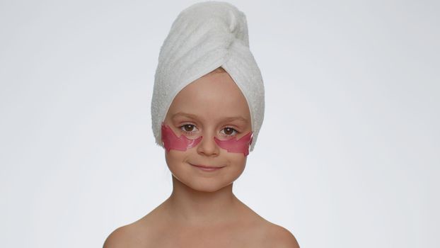 Beautiful young smiling child girl in towel on head applying pink cosmetic patches under eyes. Teenager kid face skin care treatment. Female portrait on white background. Perfect natural fresh clean