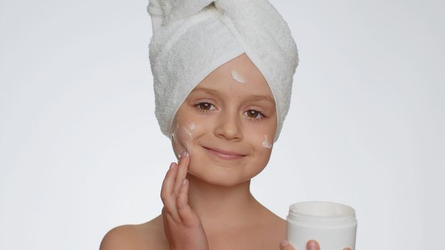 Lovely young child girl with towel on head applying cleansing moisturizing cream from creme jar. Teenager kid face skincare healthy treatment, natural cosmetics. Female portrait. Perfect fresh clean