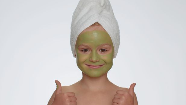 Beautiful young smiling child girl in towel on head applying cleansing moisturizing green mask on face, showing thumbs up. Teenager kid face skin care treatment, natural cosmetics. Female portrait