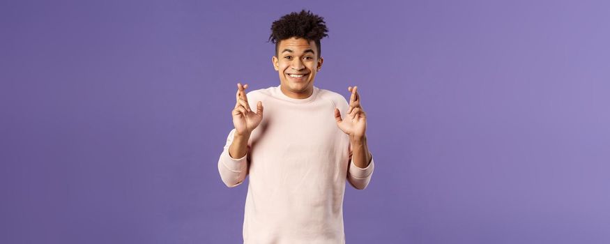 Portrait of hopeful positive, smiling young man having faith in dreams may come true, make wish, cross fingers good luck and awaiting for miracle, standing purple background praying.