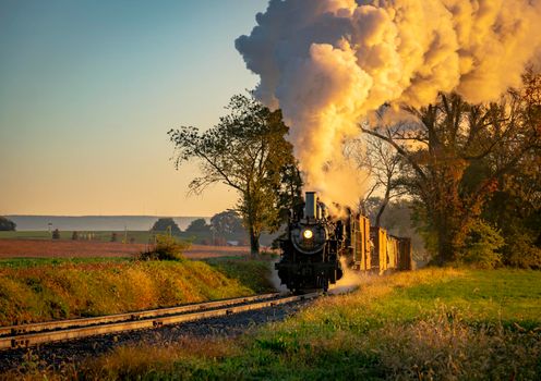 A View of an Antique Freight Steam Train Blowing Smoke Approaching Thru Trees in Late Afternoon