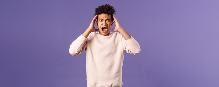 Portrait of scared and anxious young hispanic handsome man in panic, shaking hands nead head, screaming and gasping afraid, being in trouble, standing alarmed over purple background.