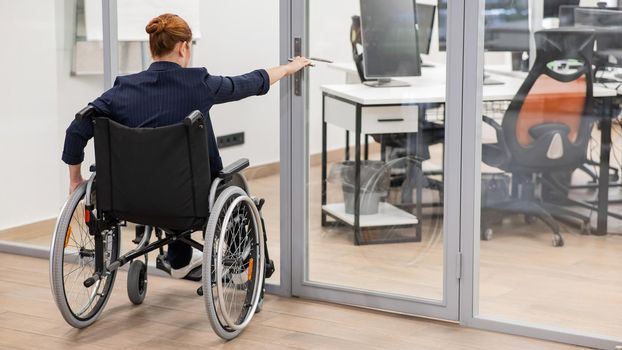 Red-haired caucasian woman in a wheelchair trying to open the door in the office