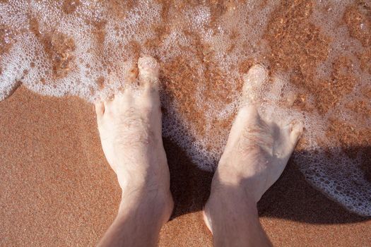 Top view of men's legs in the sea. A man is standing on the sand. An ocean wave washes the shore. Summer holidays.