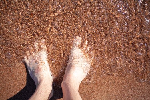 Top view of men's legs in the sea. A man is standing on the sand. An ocean wave washes the shore. Summer holidays.