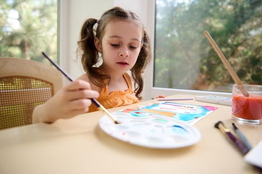 Charming Caucasian talented little girl with two ponytails, first grader sitting at a table and painting picture with watercolors and paint brush. Drawing. Art class.