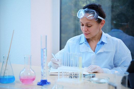 Confident multi-ethnic woman scientist, chemist-pharmacologist sits at her desk with lab glasswear and writes down the results of experiments carried out in a pharmacological laboratory in a notepad