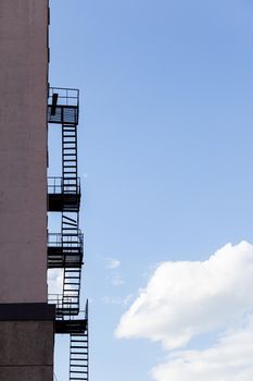 Silhouette of a fire escape on a high-rise building against a blue sky with clouds. Some of the stairs are broken. There is free space for text