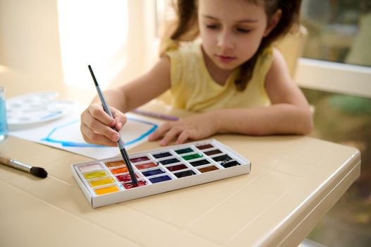 Focus on the hand of a talented little girl, holding paintbrush and mixing colorful paints on a professional watercolor palette. Learning peinture. Art class. Kids education and hobby. Back to school